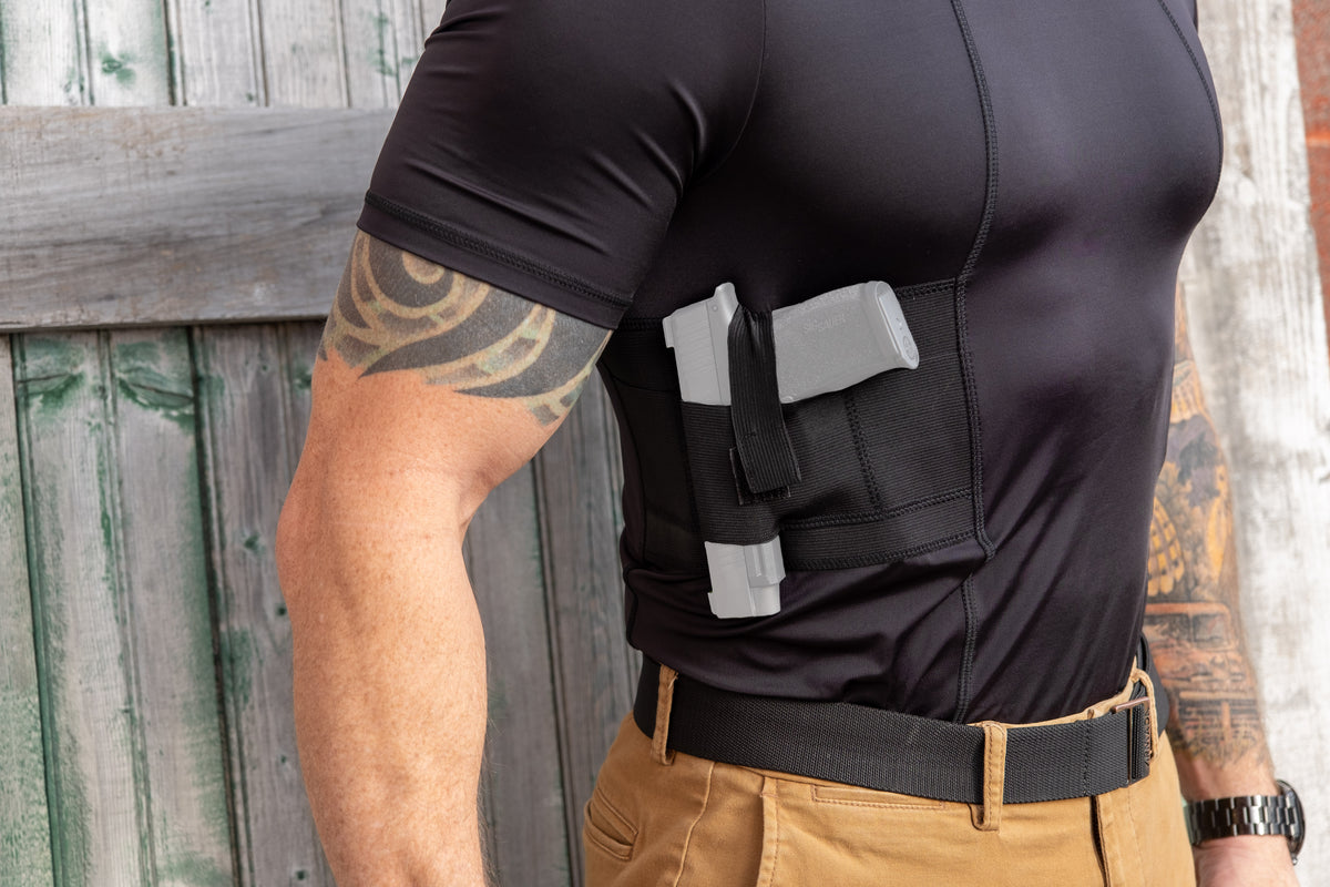 Close-up side view of a man in a black concealed carry shirt with a holstered pistol by a wooden fence