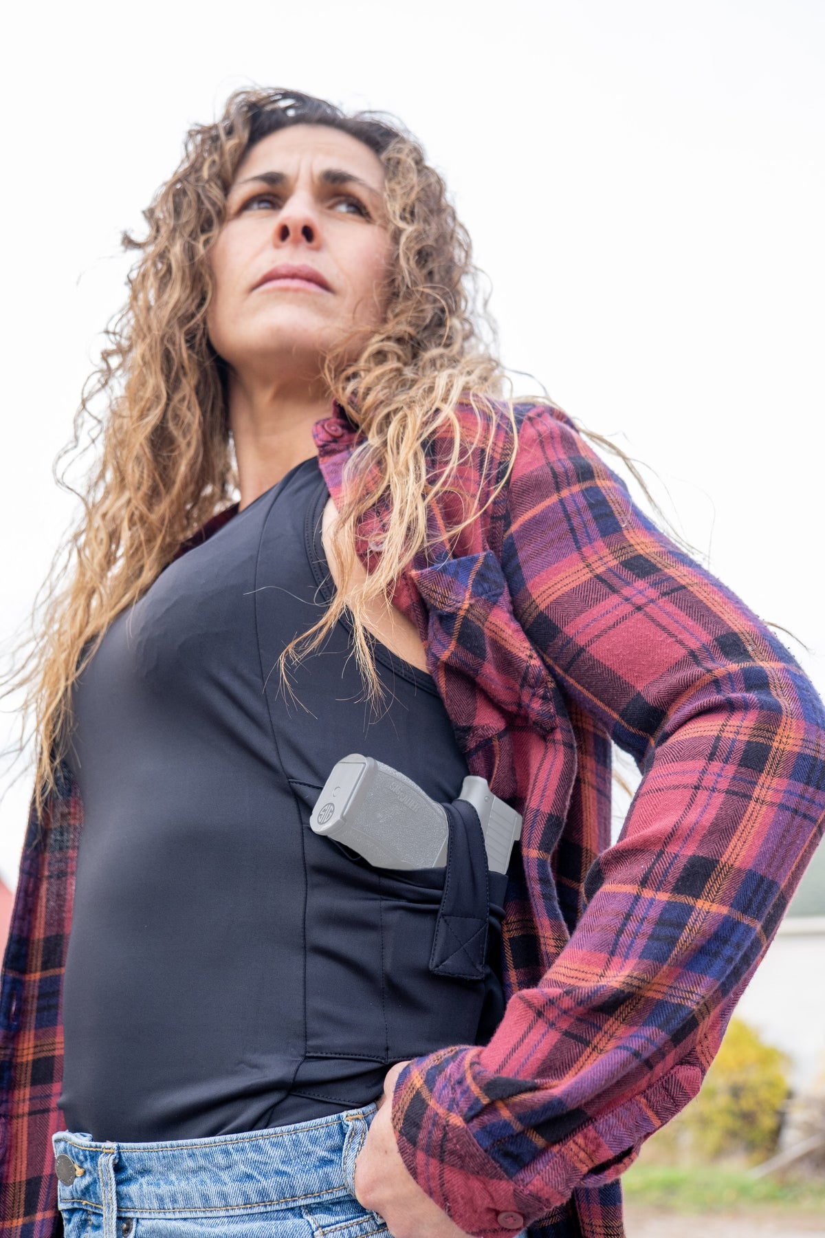 Woman wearing a black concealed carry tank top with a holstered pistol under an open plaid flannel shirt